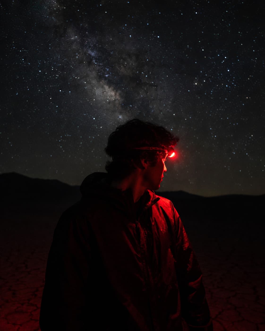 A person outdoors at night in profile, lit only by a red headlamp, the Milky Way behind them.