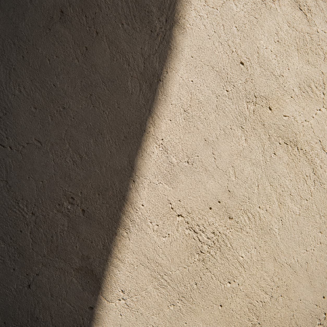 Raking light crossing a lime-plaster wall on a hard diagonal, one half in full sun and the other in shadow, trowel marks picked out along the edge.