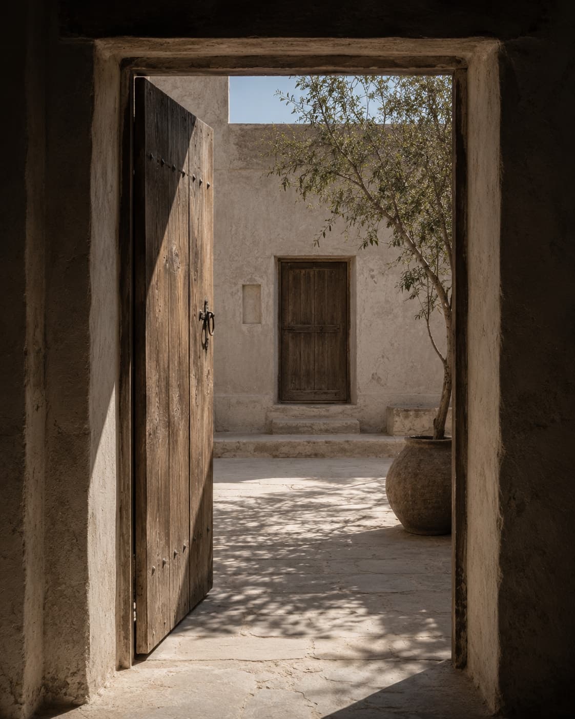 A heavy timber door standing open onto a small walled courtyard of pale flagstone, one tree against a high plaster wall, a second door opposite.
