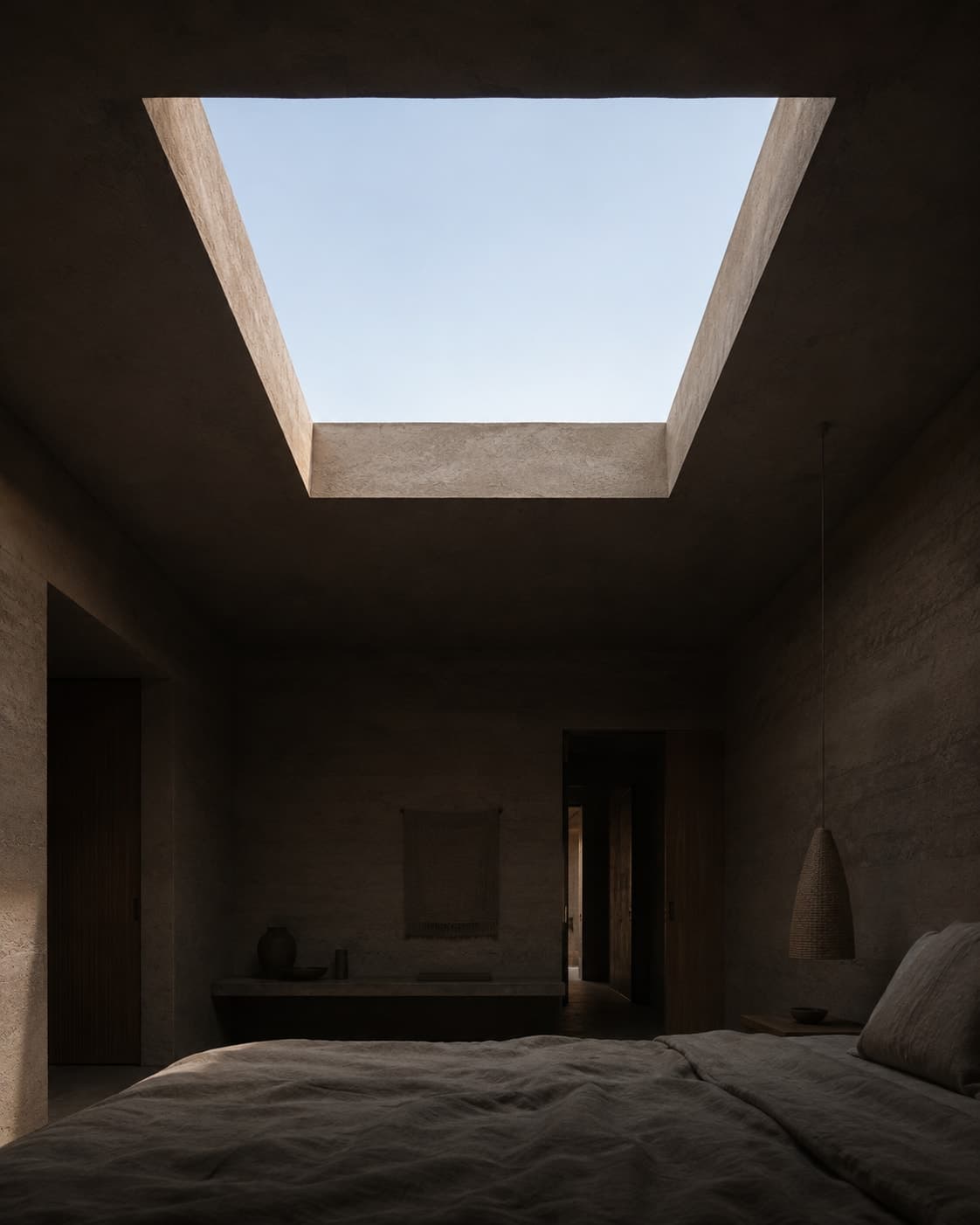 A guest room in deep shadow with a large square skylight cut flush into the ceiling directly above the bed, showing blank bright sky.